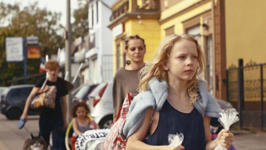 Softie film still. Young girl with family on a street, holding plastic flowers. Samuel Theis movie.