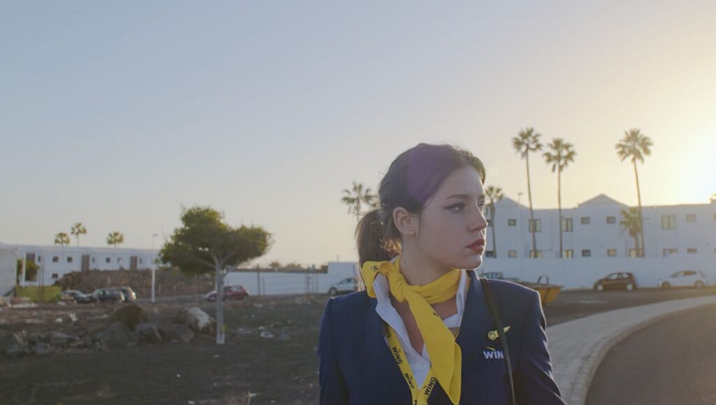 Zero Fucks Given — Emmanuel Marre & Julie Lecoustre Cannes Film Festival: Zero Fucks film still. Flight attendant in uniform outdoors. Palm trees and buildings in background.