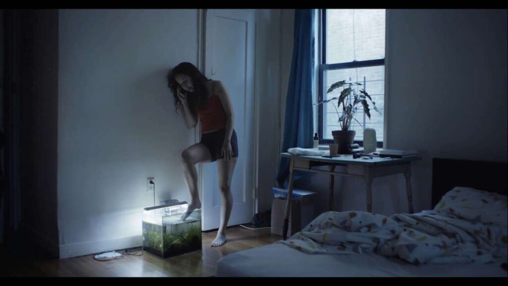 Actual People film still, Locarno Film Festival. Woman on phone with foot on aquarium in bedroom.