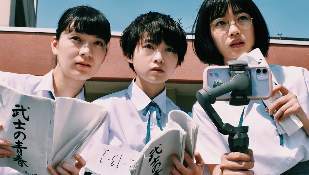 It’s A Summer Film! | Soushi Matsumoto It's a Summer Film! movie still. Three young women in school uniforms, one filming with a smartphone.