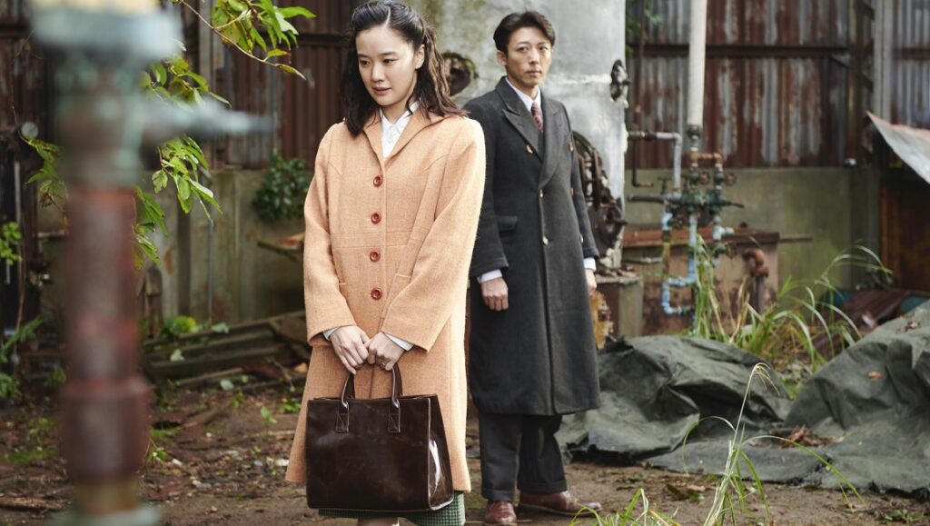 Wife of a Spy film still. Woman in coat holding bag, man in background. Kiyoshi Kurosawa movie.