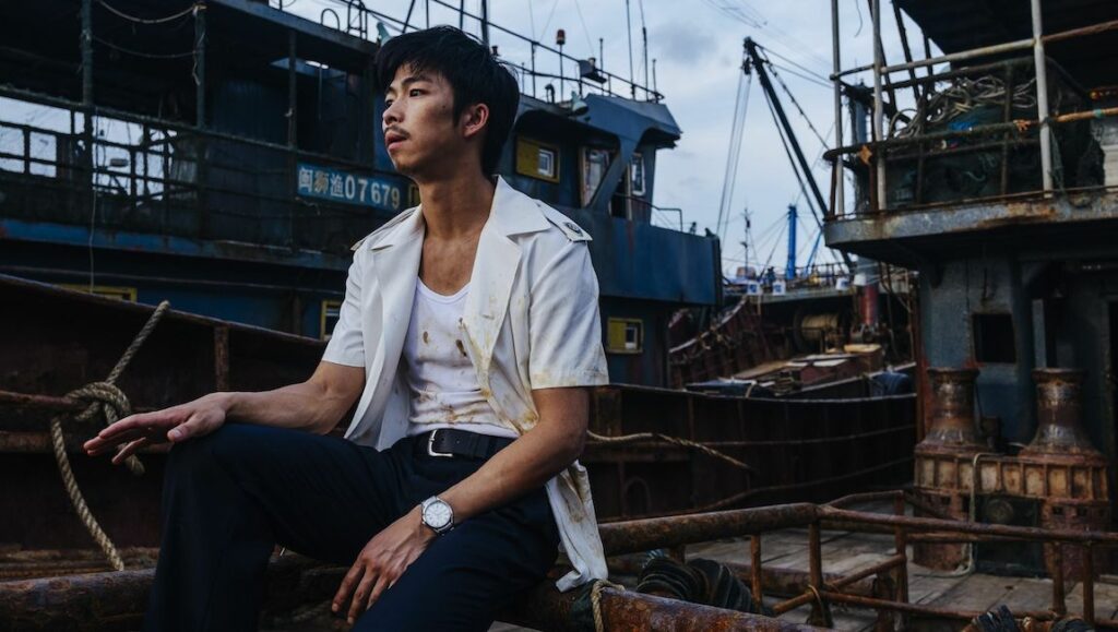 Man sits on a rusty boat at the wharf. Fishing boats in the background.
