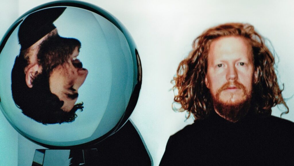 Darkside album art. Man with long red hair next to a glass ball reflecting a face. July 2021 music.