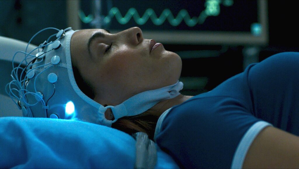 Demonic movie scene: Woman with EEG cap lying in hospital bed, brain scan monitor. Neill Blomkamp film.