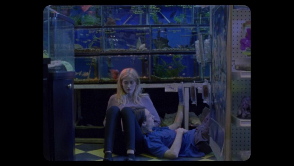 Giving Birth to a Butterfly — Theodore Schaefer Ghosting Gloria film still from Fantasia Fest 2021. Two people sit in front of fish tanks. Indie film.
