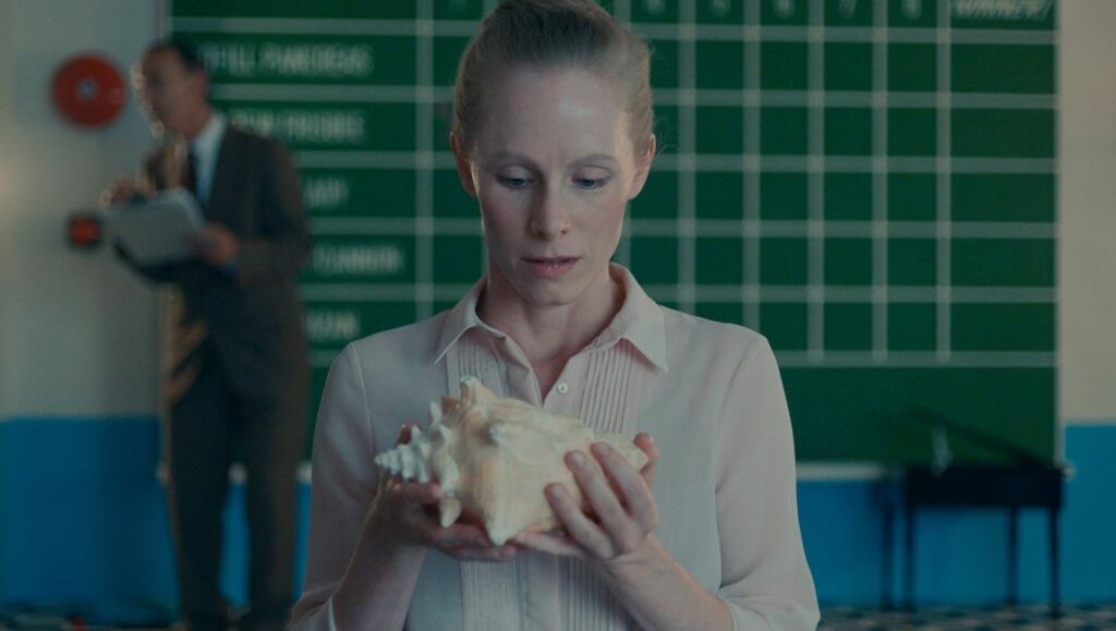 Stanleyville — Maxwell McCabe-Lokos Woman holds a seashell. Man with clipboard in background. Green grid board. Stanleyville film scene.