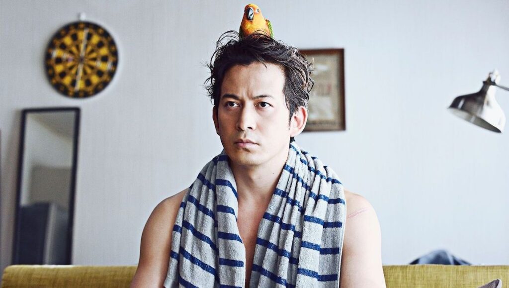 The Fable: The Killer Who Doesn’t Kill | Kan Eguchi Japanese actor with a parrot on his head. Man with towel around neck. Dartboard in background.