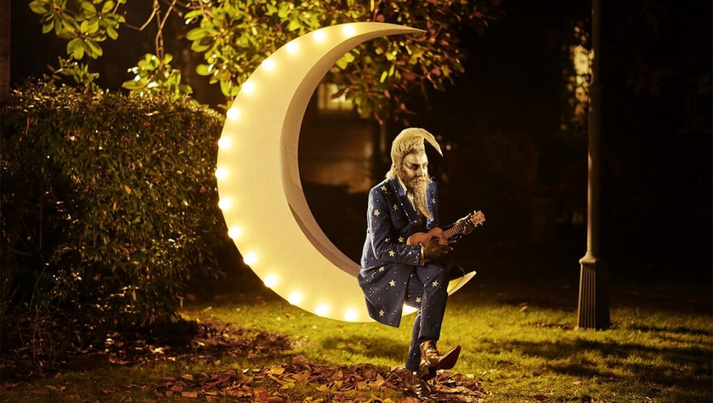 Mitch Jenkins' The Show: Man on moon playing ukulele. Night scene with illuminated crescent.