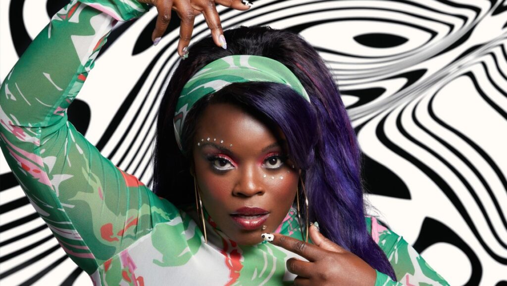Yola, soulful singer, poses in patterned green outfit. Purple-streaked hair, bold makeup, and artistic nails complete her unique look.