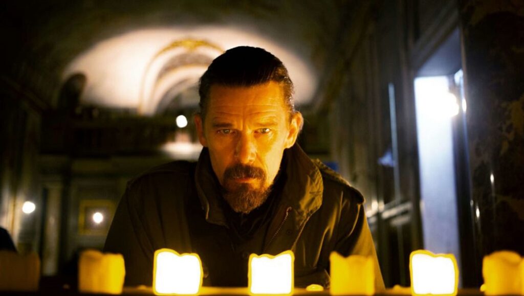 Zeros And Ones | Abel Ferrara Ethan Hawke in Zeros and Ones movie. Actor with beard stands near lit candles in a dark, arched interior.