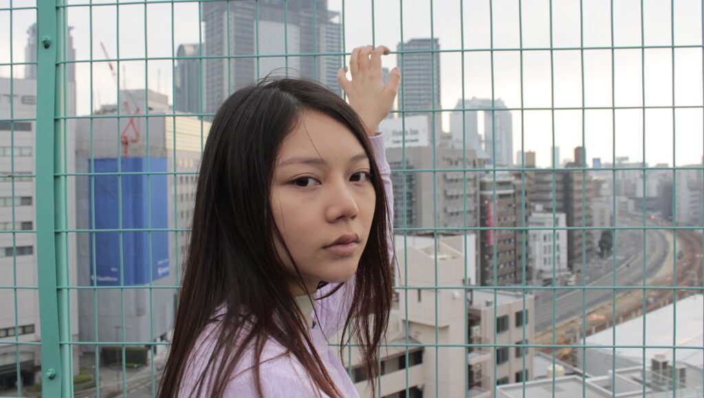 Japanese film Come and Go scene. Woman stands by fence with Tokyo cityscape in background. Japan Cuts 2021 film festival.