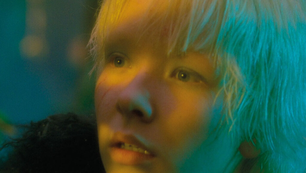 TIFF 2021: A close-up of a character from After Blue. The person has short blonde hair and is lit with yellow and blue light.