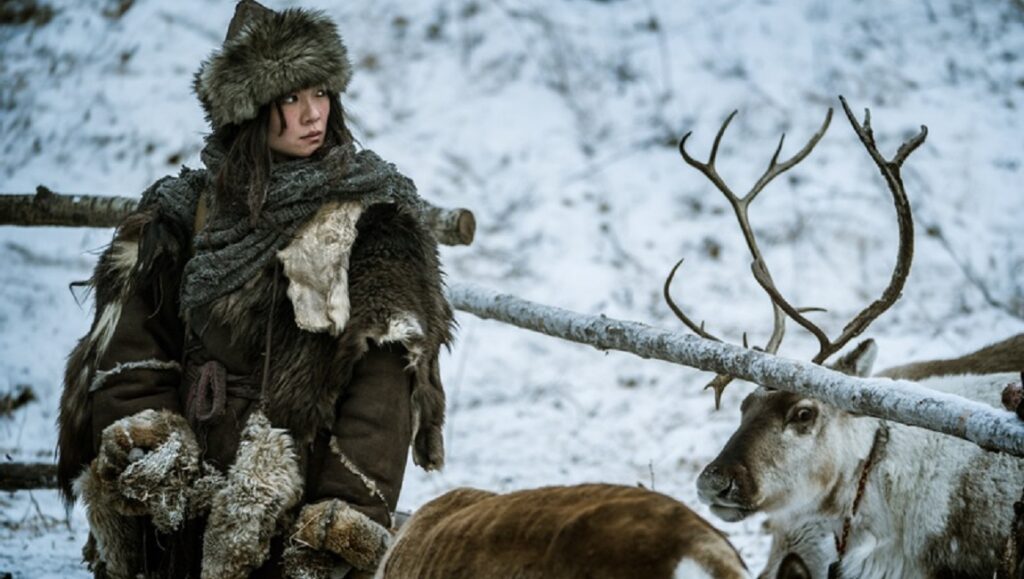 Anima | Cao Jinling Cao Jinling in Anima, wearing fur clothing and hat, standing near reindeer in a snowy landscape.