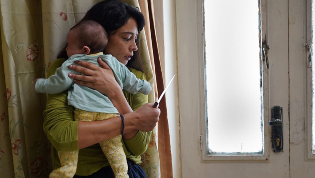 Huda’s Salon — Hany Abu-Assad Huda's Salon TIFF 2021 film still. Woman holding baby and knife, looking out window.
