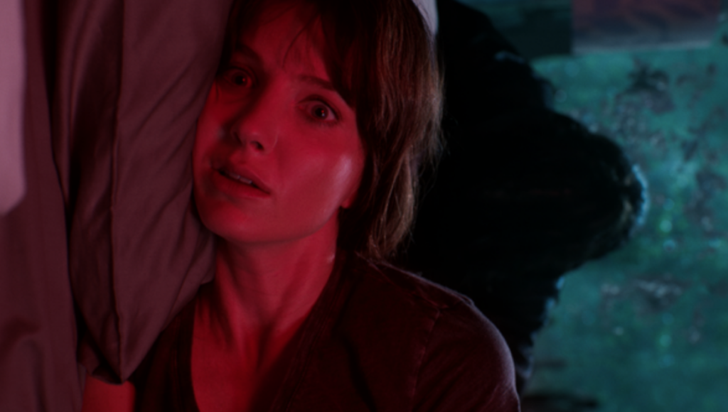 Malignant | James Wan Annabelle Wallis in Malignant, a James Wan horror film. Woman with scared expression in red lighting.