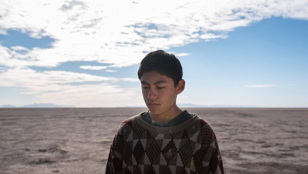 The Box movie still. A young man stands on a vast, dry plain under a bright, cloudy sky.