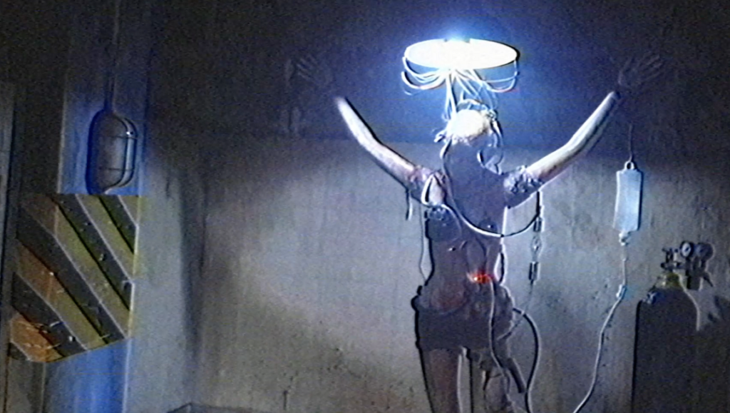 V/H/S/94 horror film still. Person wired to a light fixture, arms raised in a dark room. Gore, 90s aesthetic.