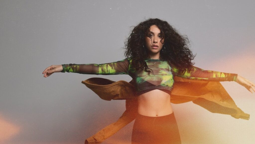 Alessia Cara | In the Meantime Alessia Cara portrait. Curly hair, green top, and brown jacket. Singer and songwriter.