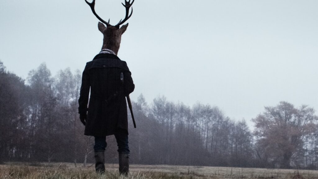 Anonymous Animals: Man with deer head in coat, standing in field. Baptiste Rouveure art.