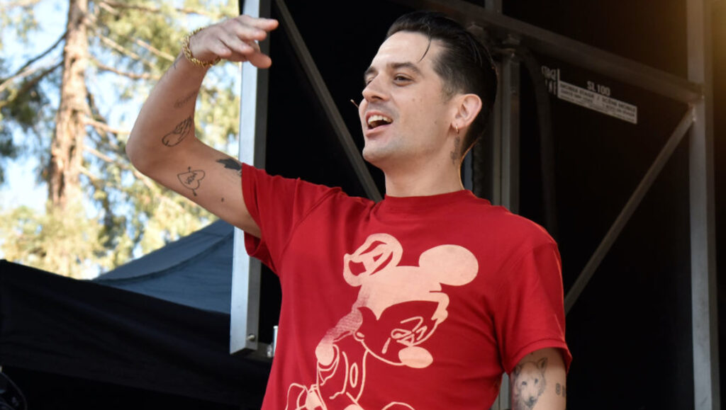 G-Eazy performing live. Rapper with tattoos, wearing a red Mickey Mouse t-shirt on stage. Concert photography.