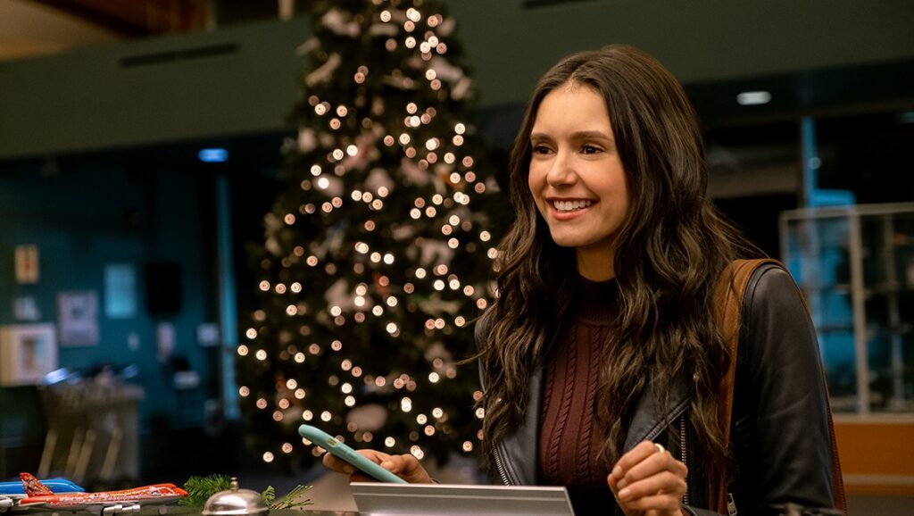 Love Hard movie scene: Nina Dobrev smiles at a hotel desk with a Christmas tree in the background.