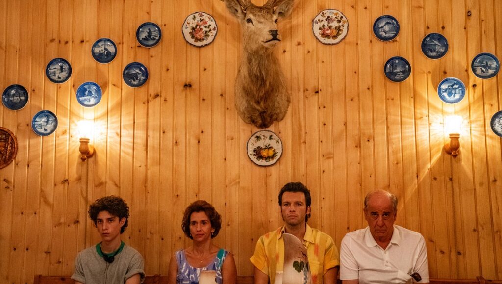 The Hand of God scene: Family sits below deer head trophy and plates on wood wall. Paolo Sorrentino film.