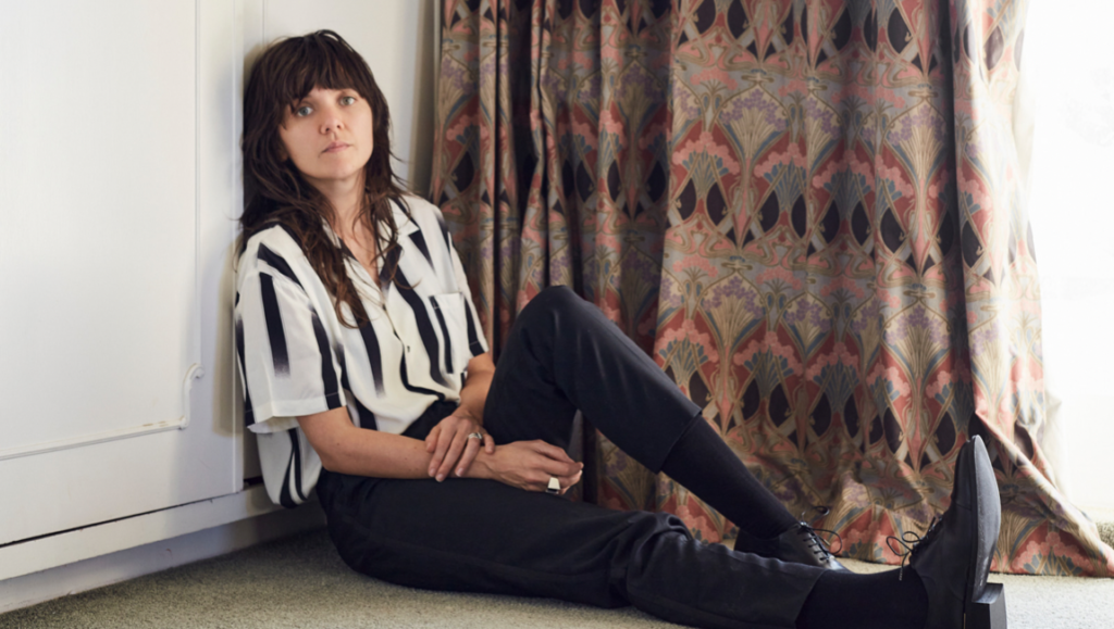 Courtney Barnett, November 2021 album artist. Indie rock musician in striped shirt and black pants sitting on the floor.