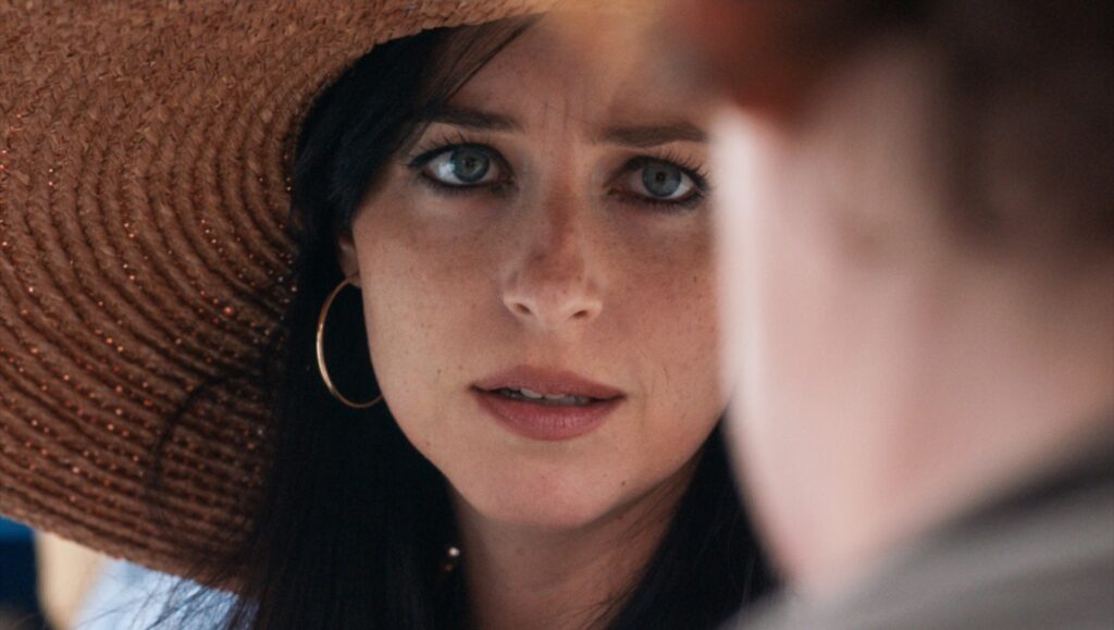 The Lost Daughter | Maggie Gyllenhaal Dakota Johnson in The Lost Daughter. Close-up of her face with a straw hat and hoop earrings.