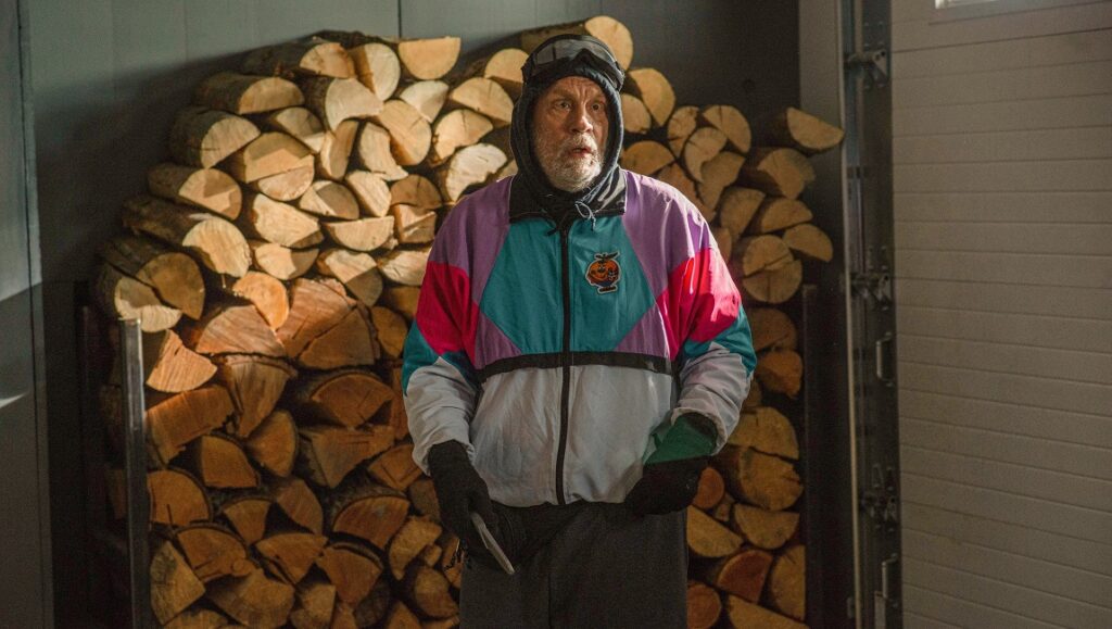 Shattered — Luis Prieto Still from Shattered (2022) featuring John Malkovich in a colorful jacket, standing near a wood pile.