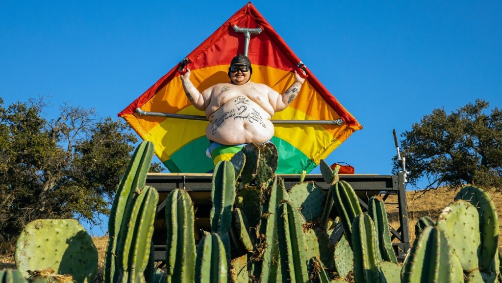 Jackass Forever — Jeff Tremaine Jackass Forever: Man with rainbow kite and tattoos stands behind cacti.