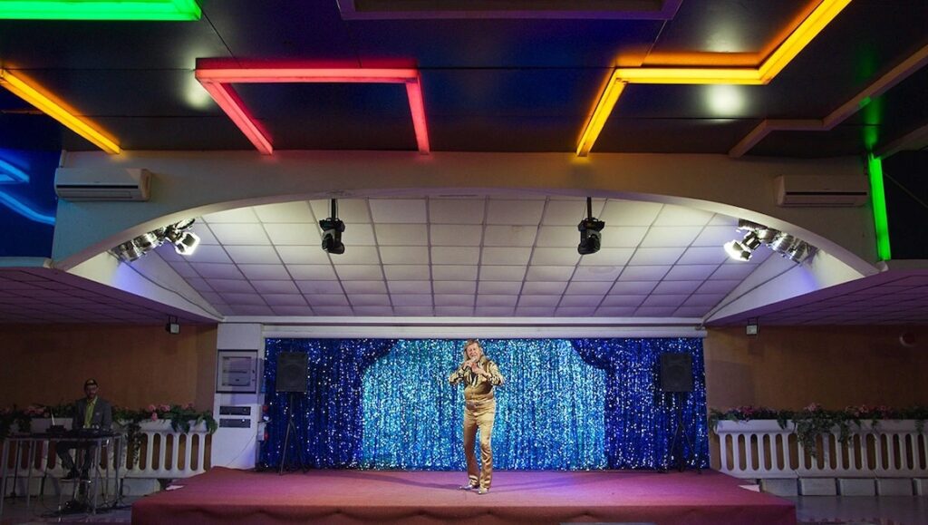 Still from Rimini at the Berlin Film Festival 2022. Singer in gold suit on stage with blue curtain.