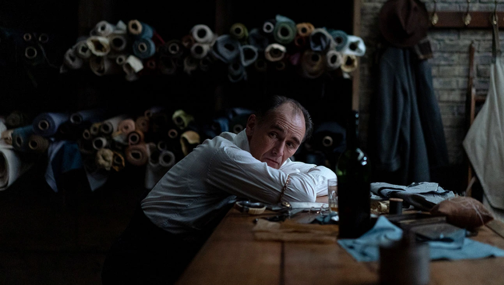 The Outfit — Graham Moore Mark Rylance in The Outfit movie. Tailor rests at his workbench surrounded by rolls of fabric.