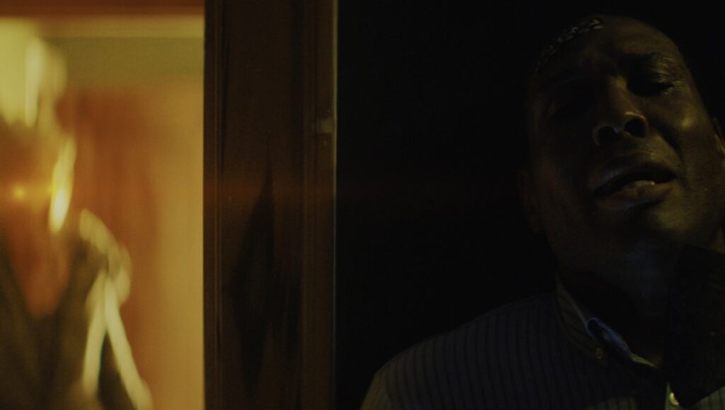 Night’s End — Jennifer Reeder Still from Jennifer Reeder's Night's End horror film. A man looks on in fear as a shadowy figure lurks in the background.