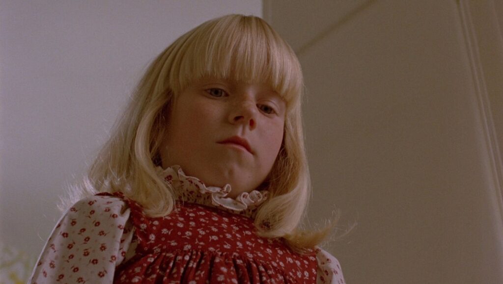 Still from The Brood: A young blonde girl in a red floral dress looks down. Cronenberg horror film.
