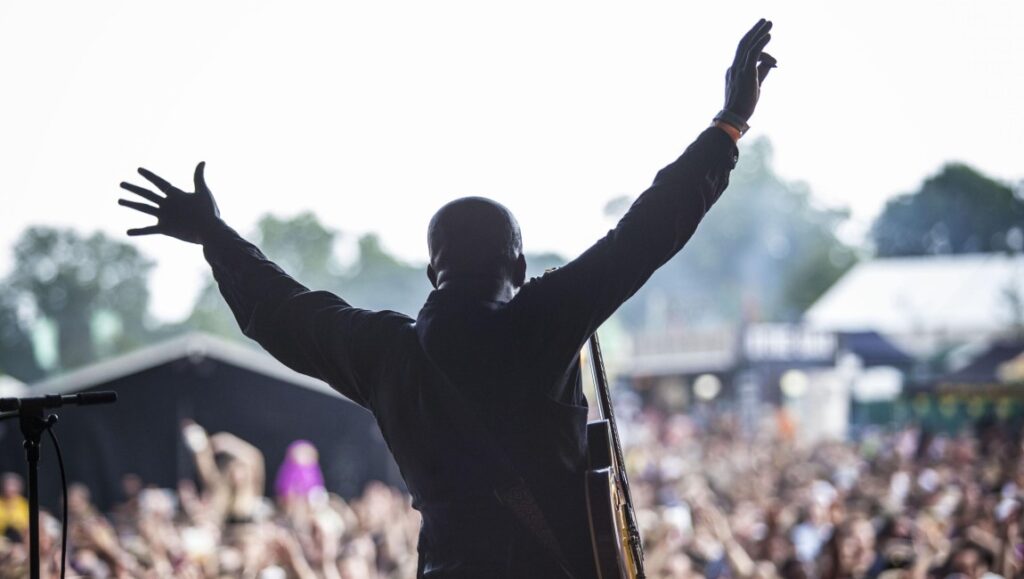 Cymande musician performing live at music festival. Silhouette of artist with arms raised in front of concert crowd.