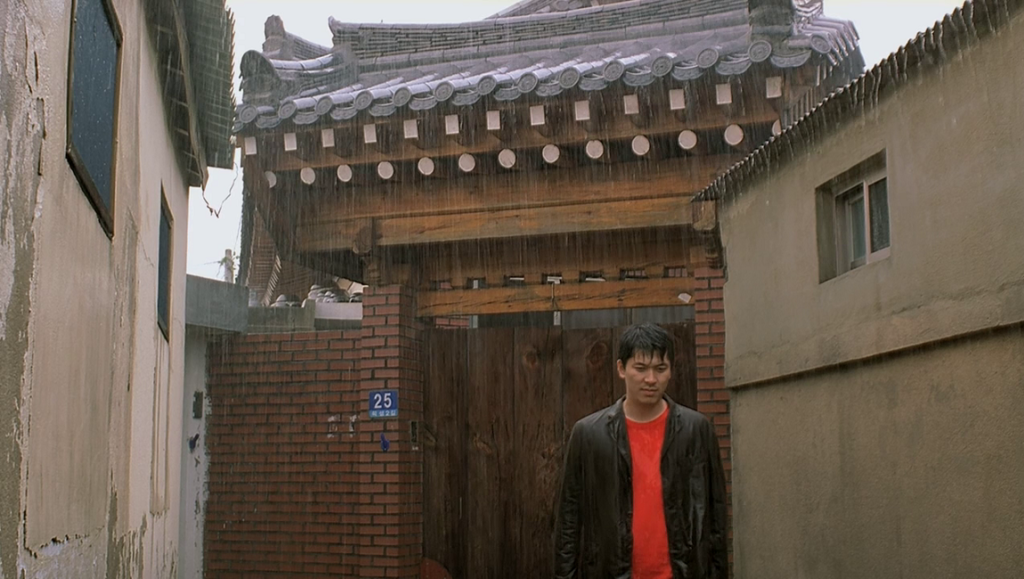 On the Occasion of Remembering the Turning Gate film still. Man in red shirt, traditional Korean architecture in rain.