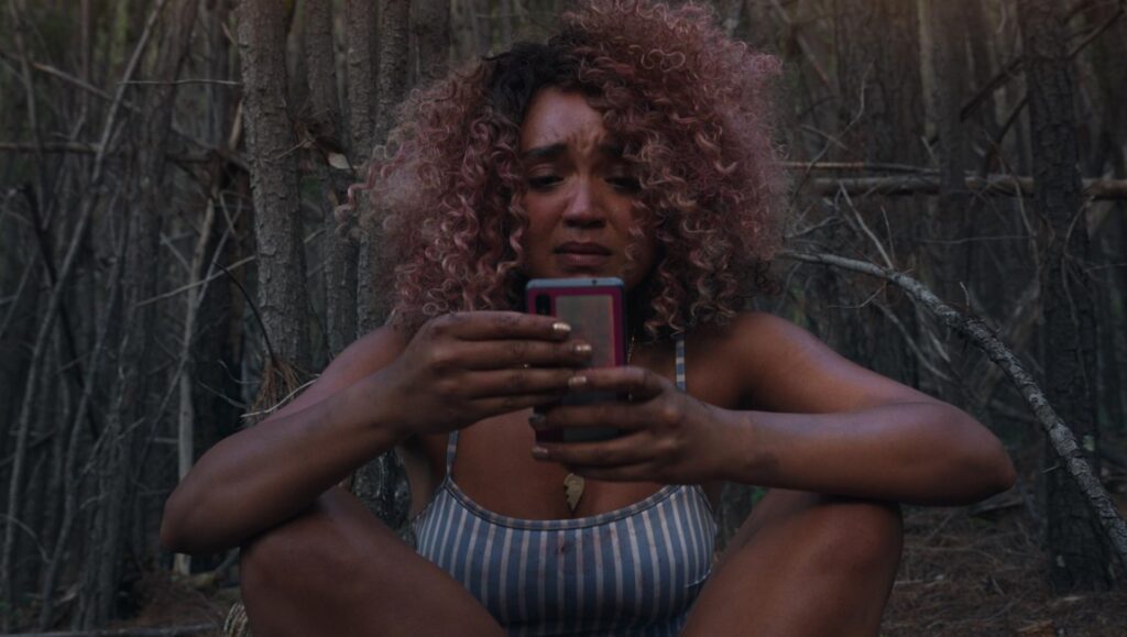 Woman with curly hair looks at phone with worried expression. Dark background.