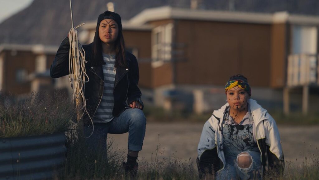 Two Indigenous teens in Slash/Back movie. Girl holding rope, wearing black jacket. Girl in overalls with face paint.