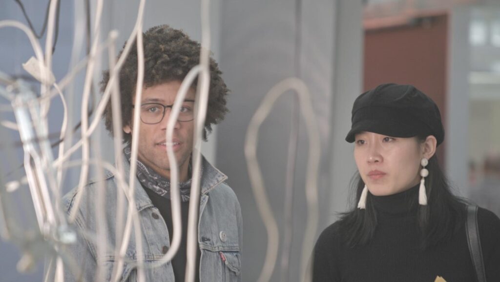 Two people at an art gallery. Man with glasses, woman with hat and earrings. Art installation in foreground.