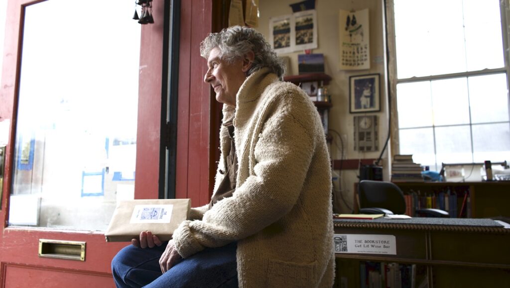 Matthew Tannenbaum in Hello Bookstore. Man sits in bookstore holding book. Books, calendar, and desk visible.