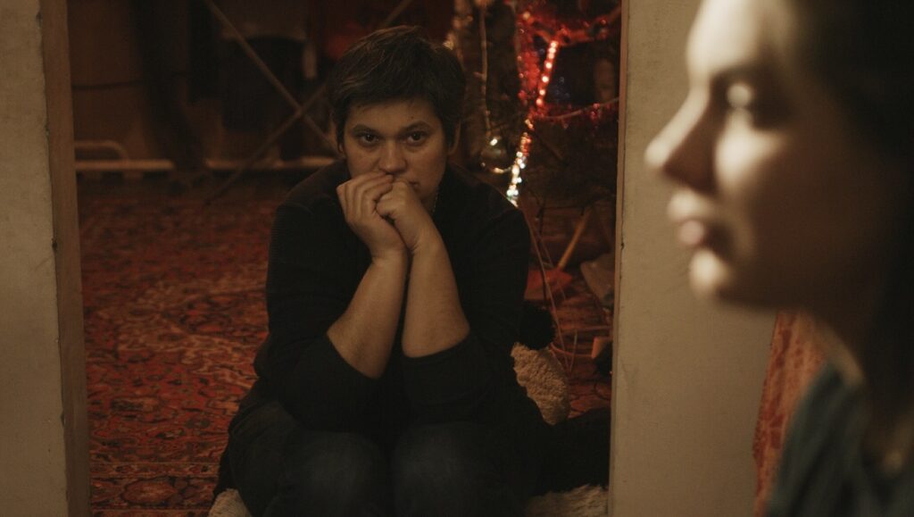 Indoor scene with two women. One woman is seated, looking pensive. The other woman is out of focus.