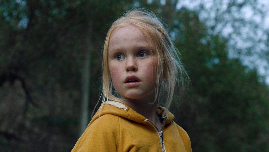Young girl in The Innocents movie. Blonde hair, yellow hoodie, looking off-camera with a slightly surprised expression.