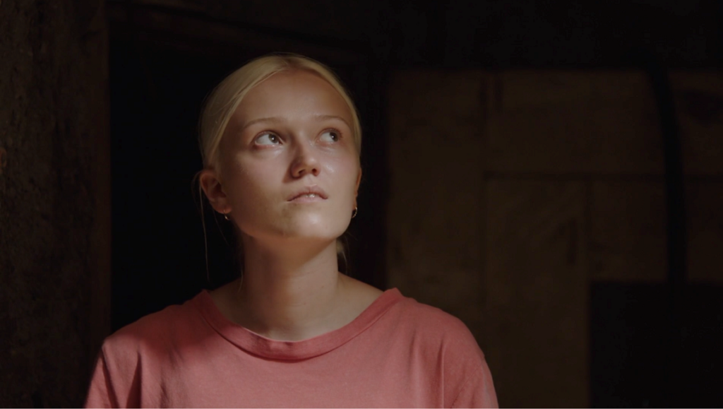 Still from Pilgrims movie. Young woman with blond hair looks up in dim light. Laurynas Bareisa film review.