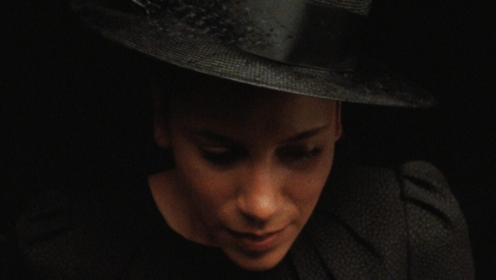 Geographies of Solitude film still. Woman in hat, dark background. Art of the Real 2022.