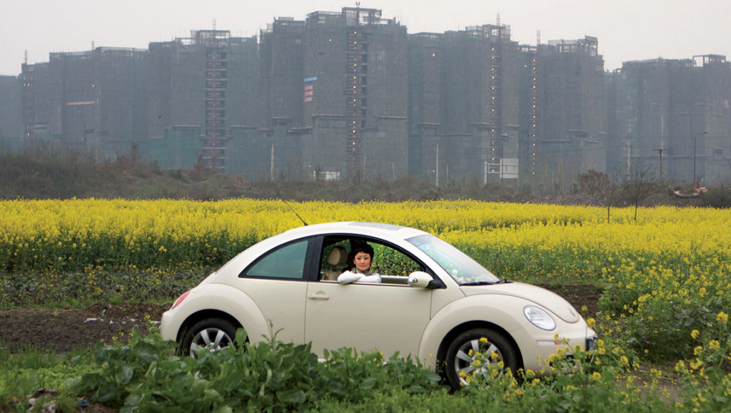 Jia Zhangke's 24 City scene: Woman in a VW Beetle amidst yellow flowers, with city buildings under construction in the background.