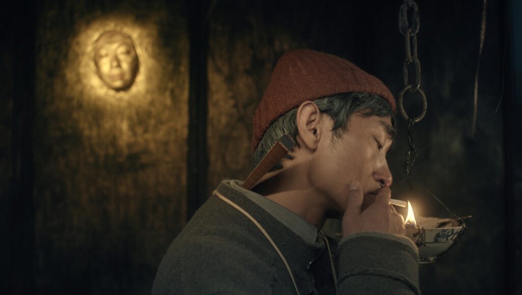 A New Old Play — Qiu Jiongjiong A New Old Play film still. Man smoking, wearing a red cap. Face sculpture on wall. Qiu Jiongjiong film.
