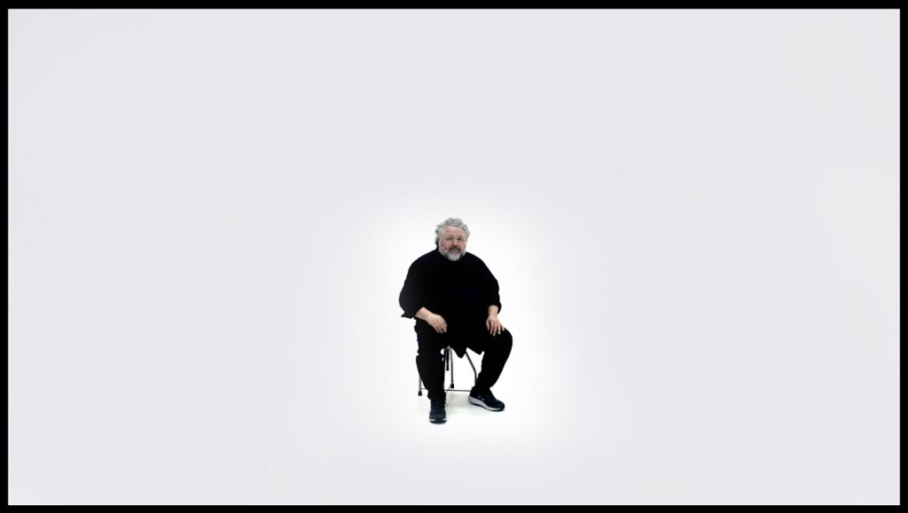 Mau, Before We Vanish. Man with white hair wearing black, seated on a stool against a white background.