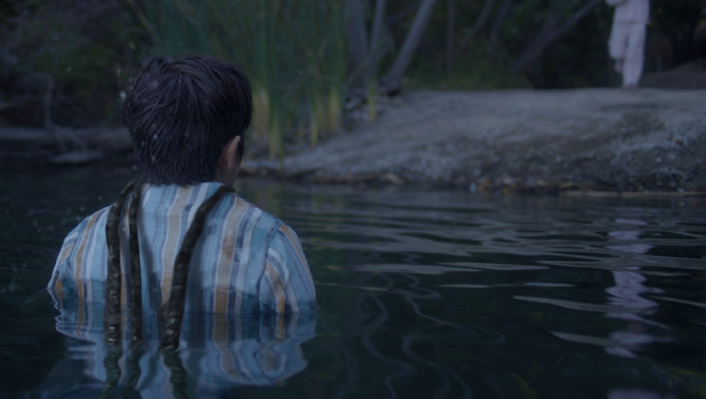 Monstrous movie still. A person stands in water, facing away from the camera, with another figure on the shore.