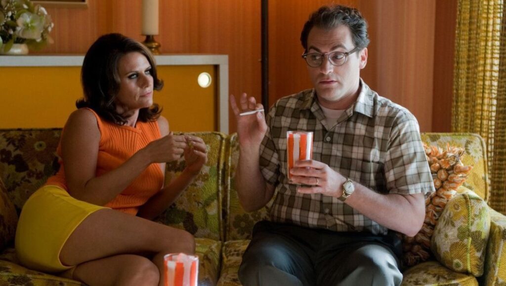 A Serious Man movie scene. Man in glasses, plaid shirt with a woman in orange top, yellow skirt on floral couch.