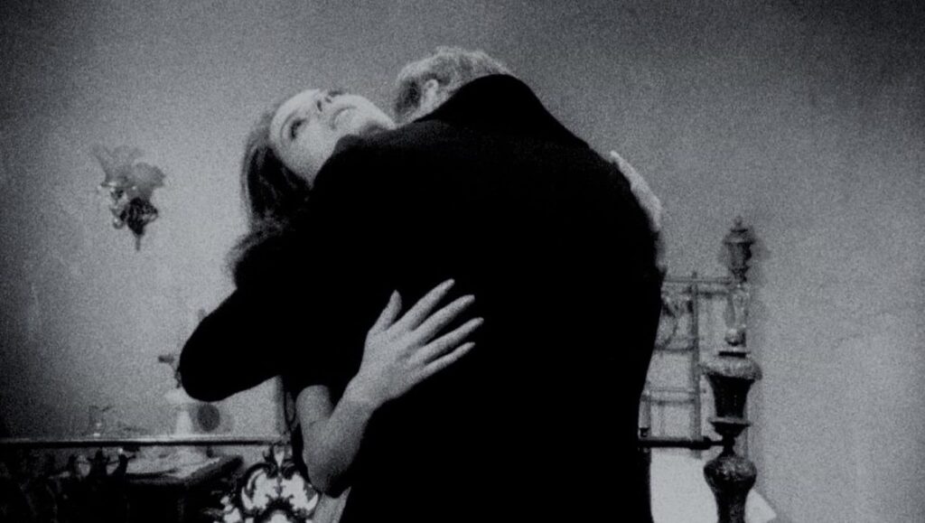 Black and white still from Cuadecuc, vampir film. Woman embraces a man; vampire theme.